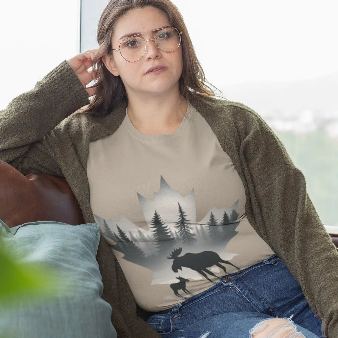 Woman wearing a beige t-shirt with a moose and forest design, sitting on a couch.